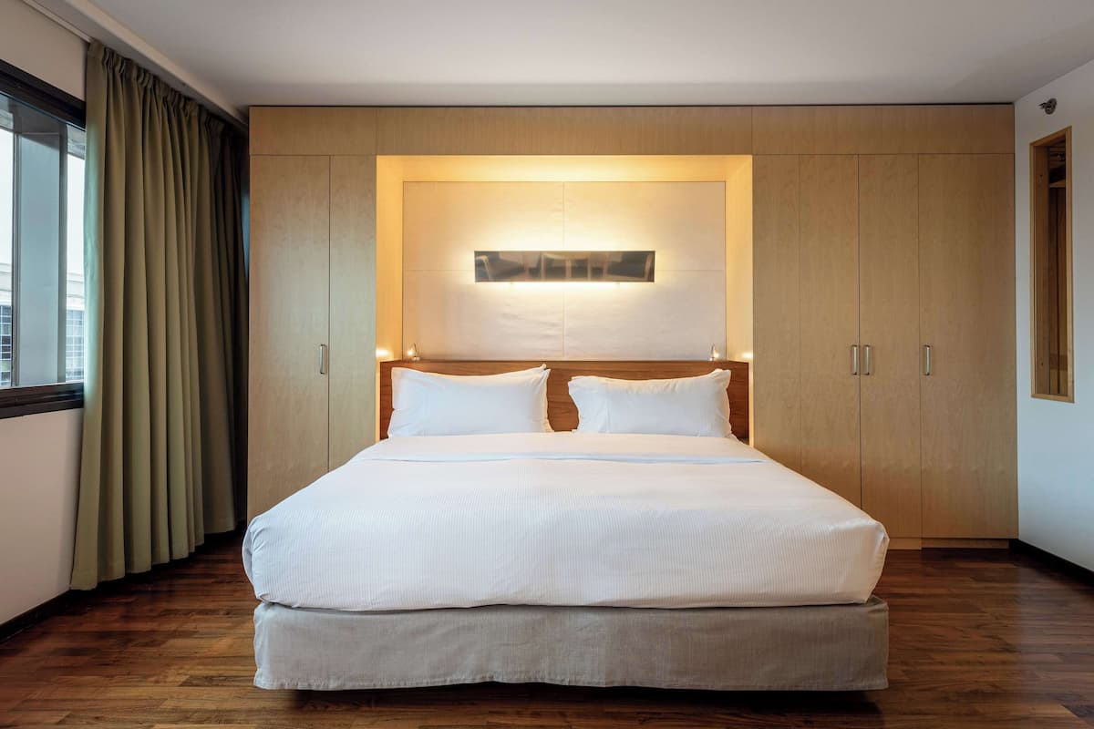 Ein ordentlich bezogenes Bett in einem Hotelzimmer mit Holzboden, einem großen Fenster mit Vorhängen und einem Holzschrank mit einer Leiste darüber.
