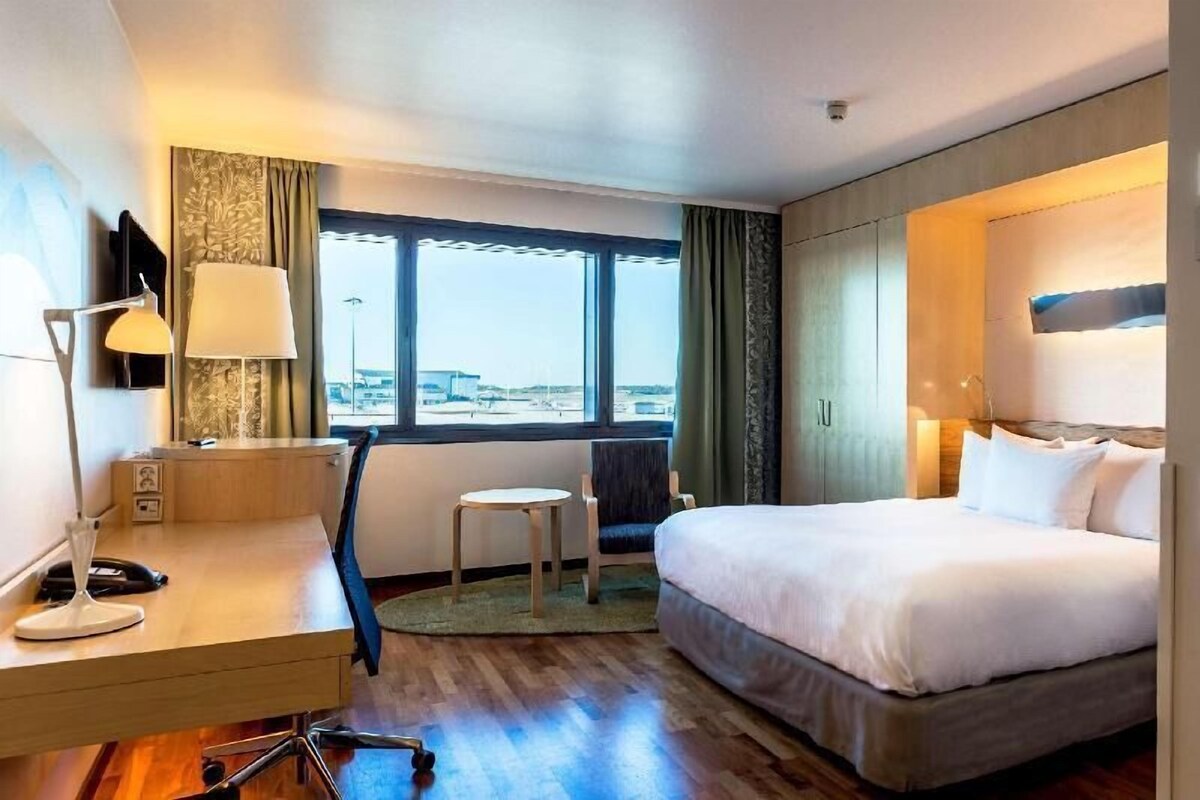 A hotel room with a bed, desk, chair, lamp, and large window.