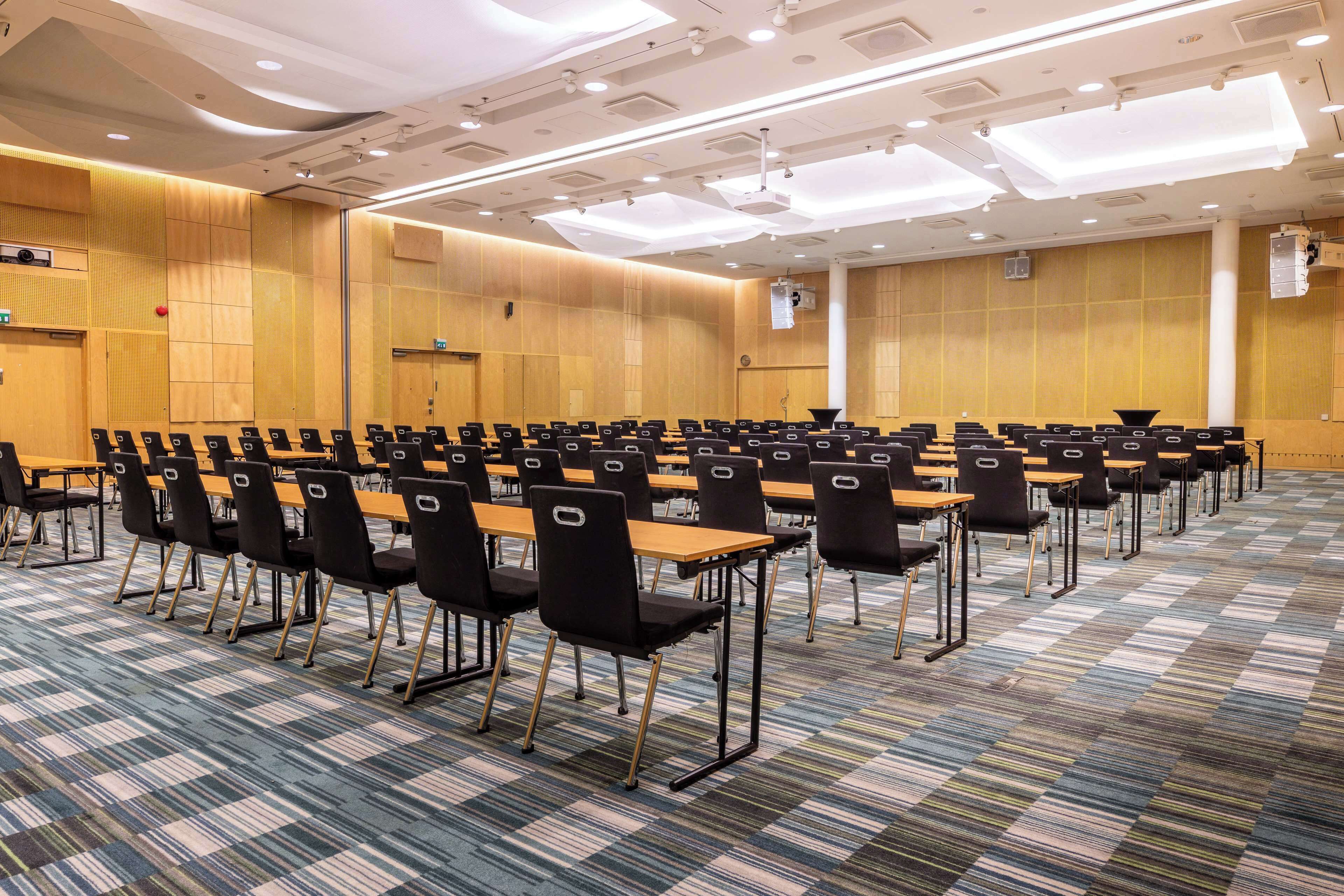 Une salle de conférence avec des rangées de tables et de chaises, une moquette à motifs et un mur en bois sur lequel est fixé un projecteur.