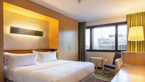 A hotel room with a large bed, a desk, a chair, a small table, and a window with a view of buildings.