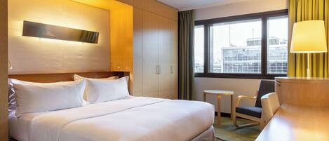 A hotel room with a large bed, a desk, a chair, a small table, and a window with a view of buildings.