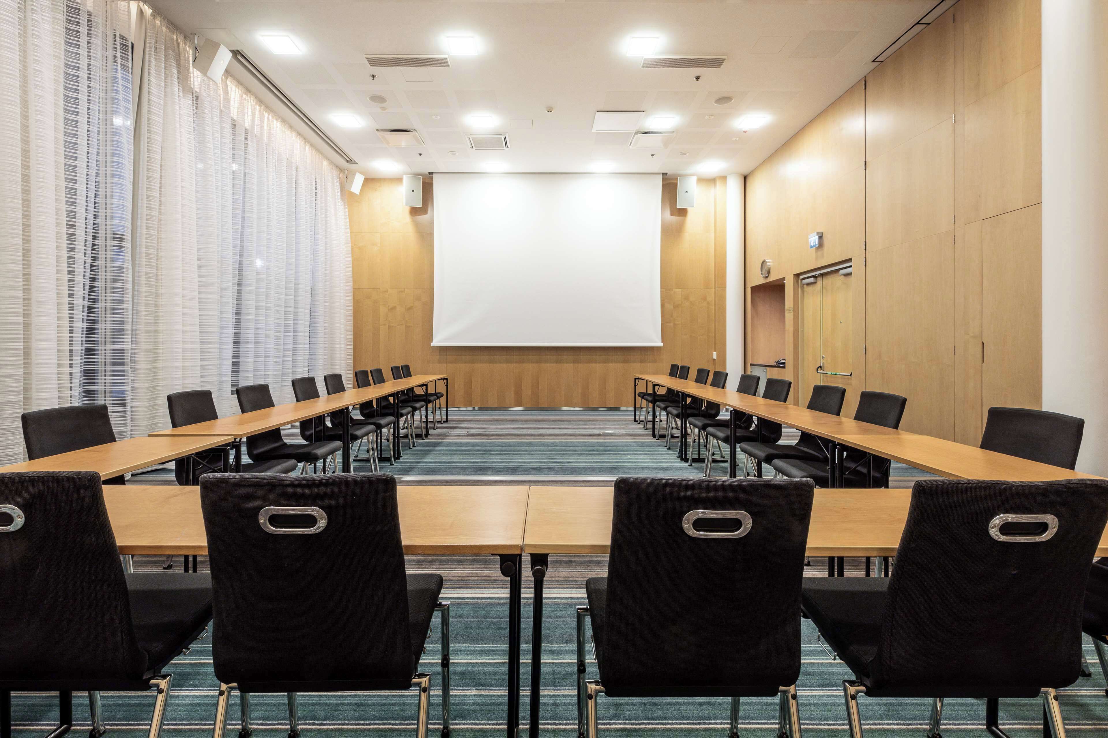 Une salle de conférence avec une longue table, des chaises noires, un écran de projection et un mur recouvert de panneaux en bois.
