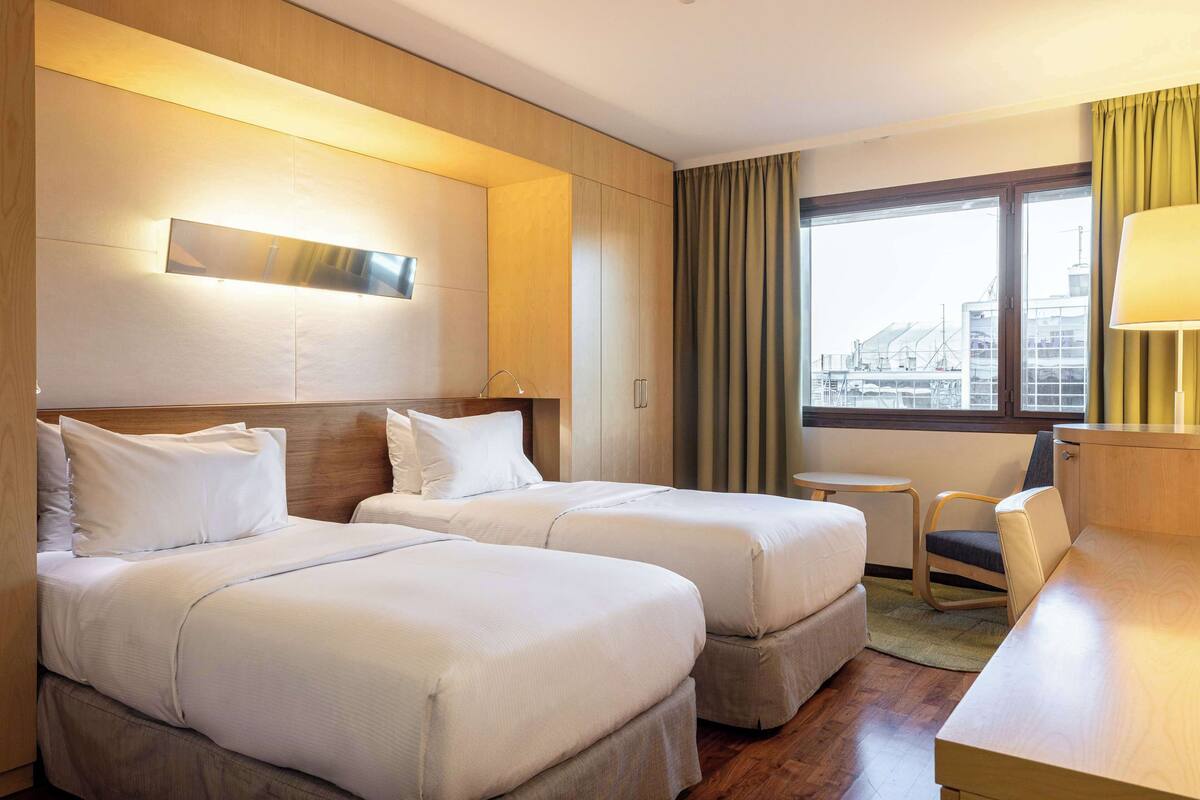 Ein Hotelzimmer mit zwei Betten, einem Schreibtisch, einem Stuhl, einem kleinen Tisch und einem Fenster mit Vorhängen.