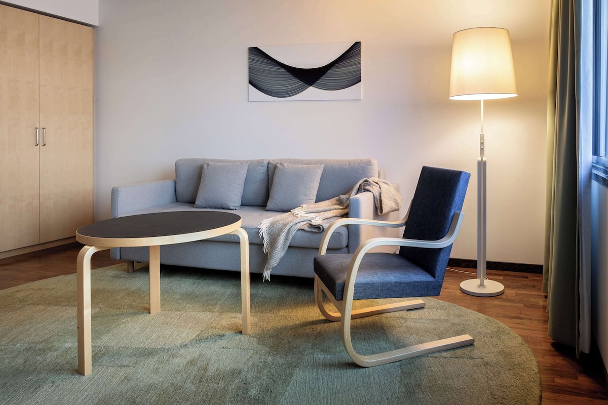 Ein modernes Hotelzimmer mit grauem Sofa, einem kleinen runden Tisch, einem blauen Sessel, einer Stehlampe und einem abstrakten Kunstwerk an der Wand.