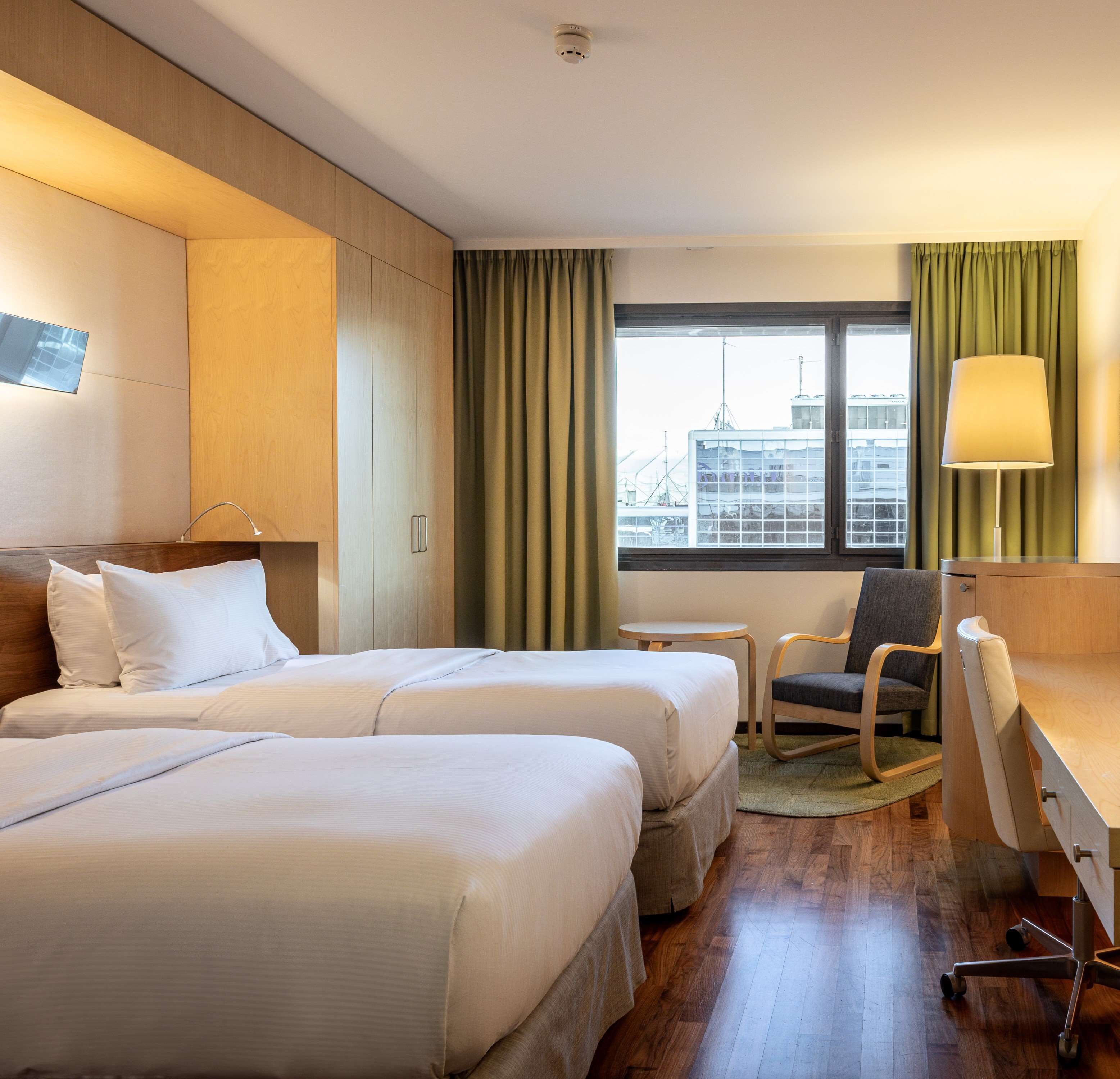 A hotel room with two beds, a desk, a chair, a lamp, and a window with a view of buildings.
