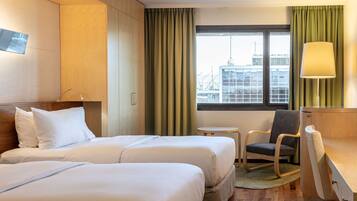 A hotel room with two beds, a desk, a chair, a lamp, and a window with a view of buildings.