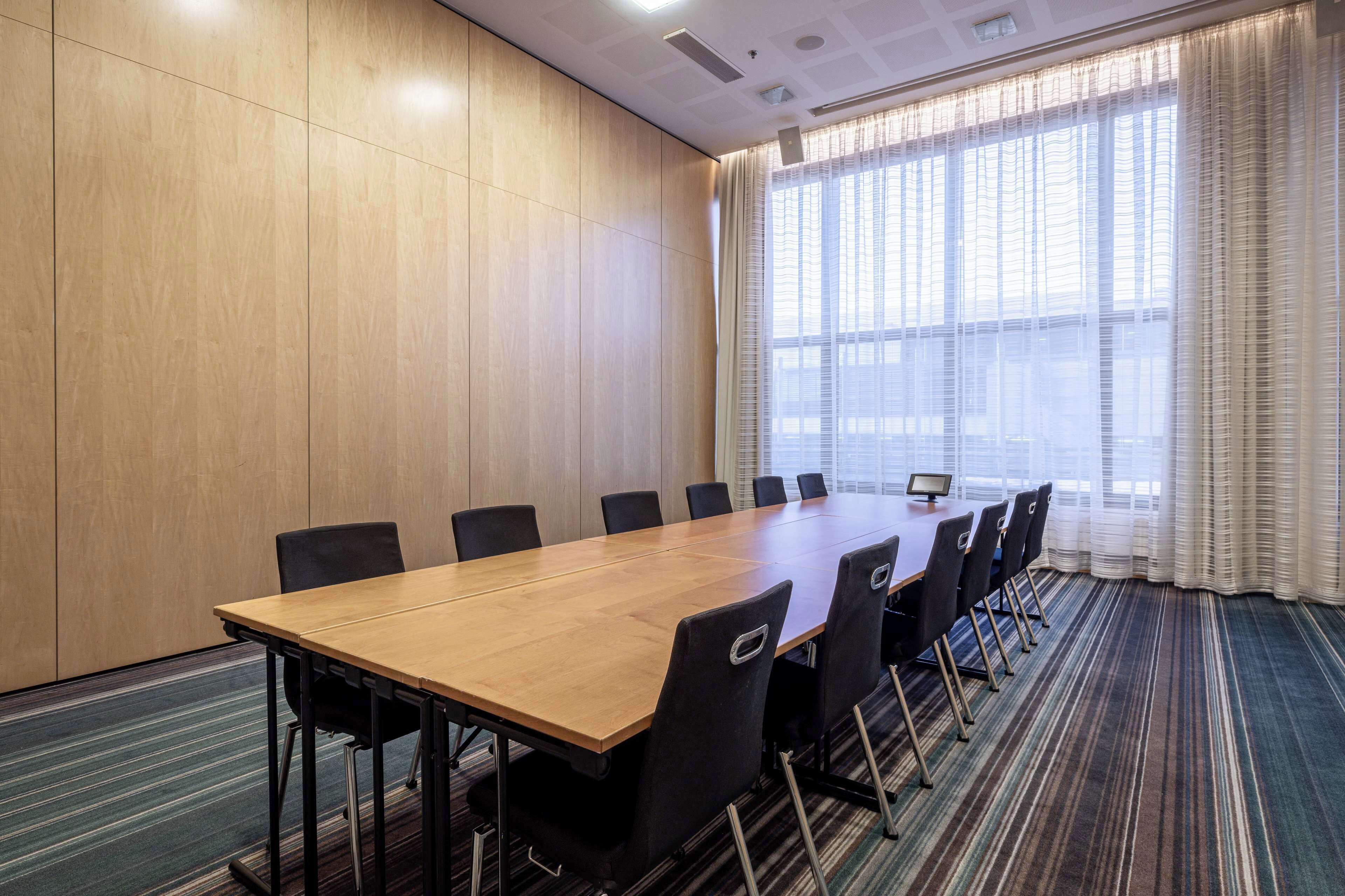 Uma sala de conferências com uma mesa comprida, cadeiras pretas e uma janela grande com cortinas.
