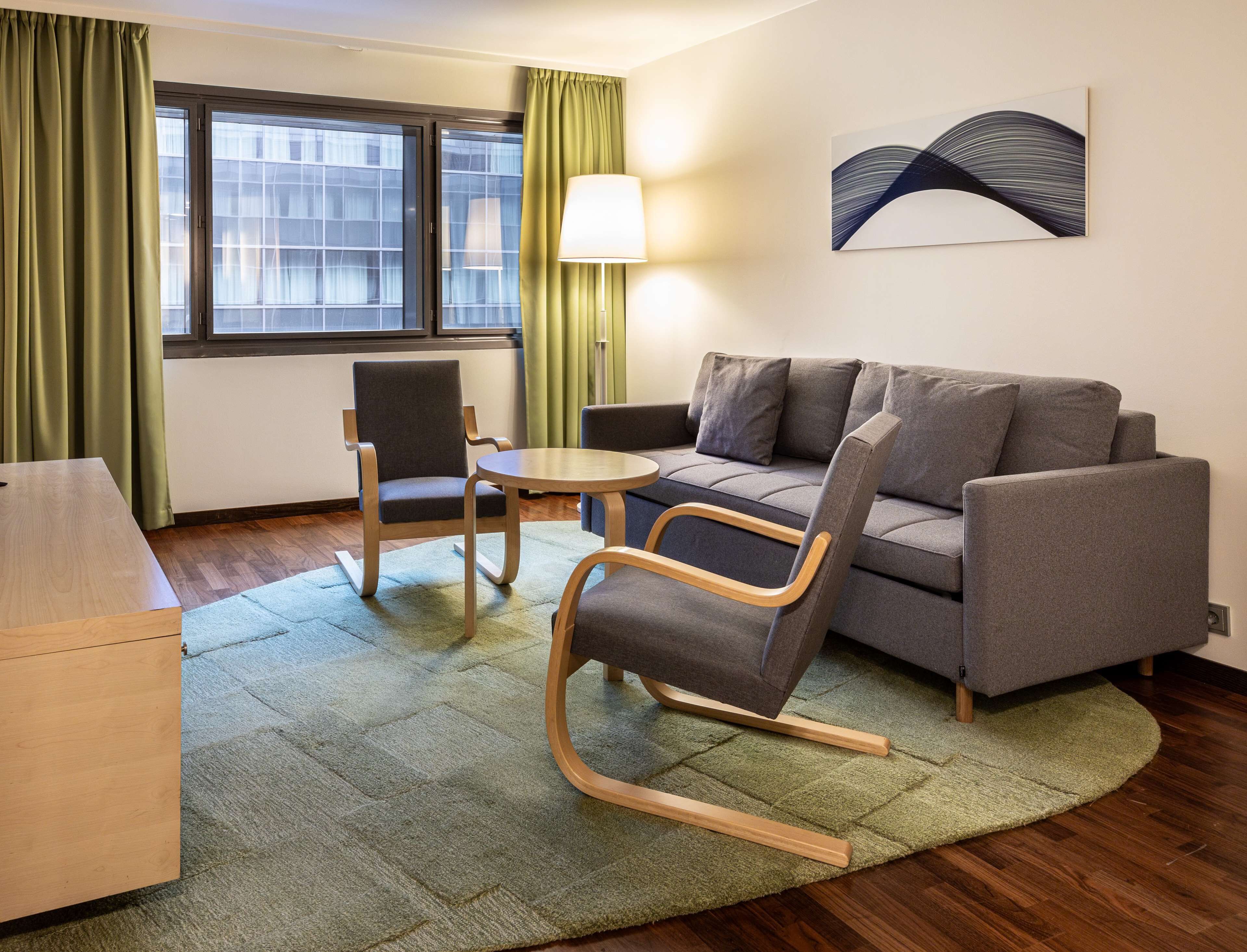 Une chambre d’hôtel moderne avec un canapé gris, une chaise en bois, une petite table ronde et un tapis vert.