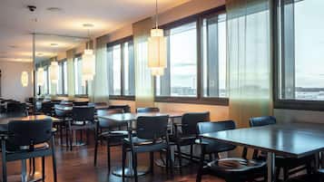 Uma sala de jantar moderna com várias mesas e cadeiras, janelas amplas com cortinas e iluminação pendente.