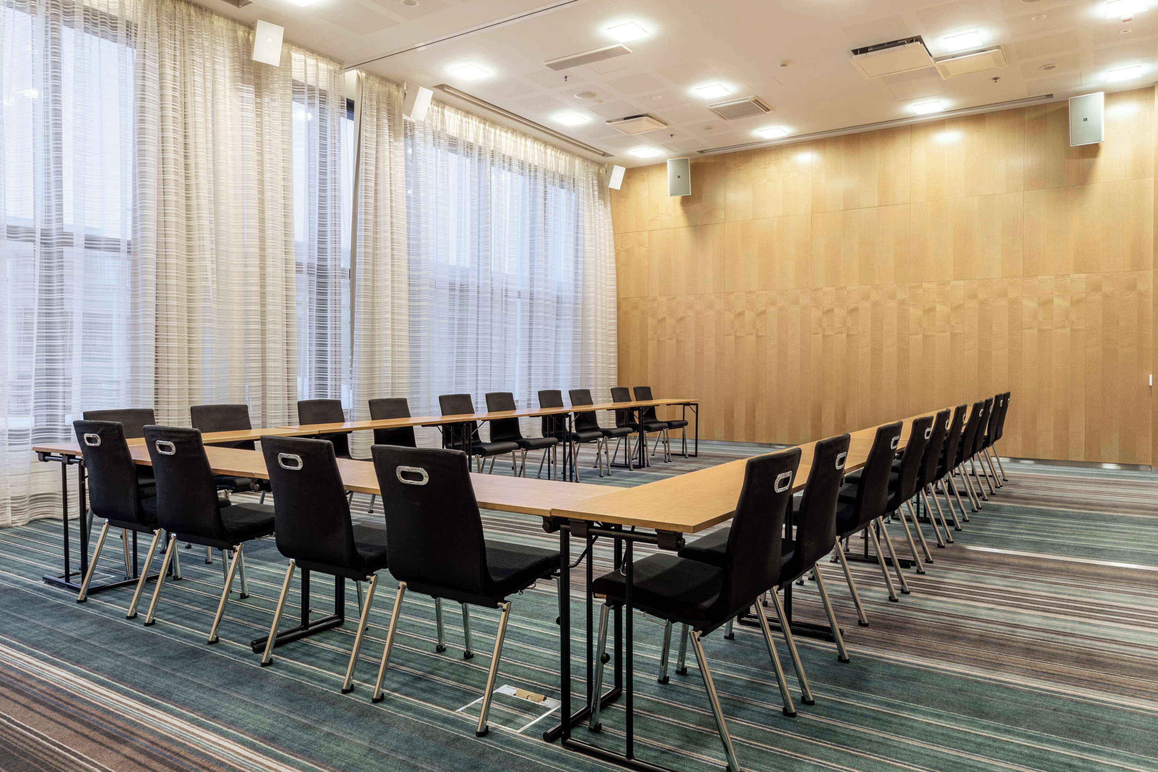 Une salle de conférence avec une longue table et des chaises, de grandes fenêtres équipées de stores et un mur en bois.