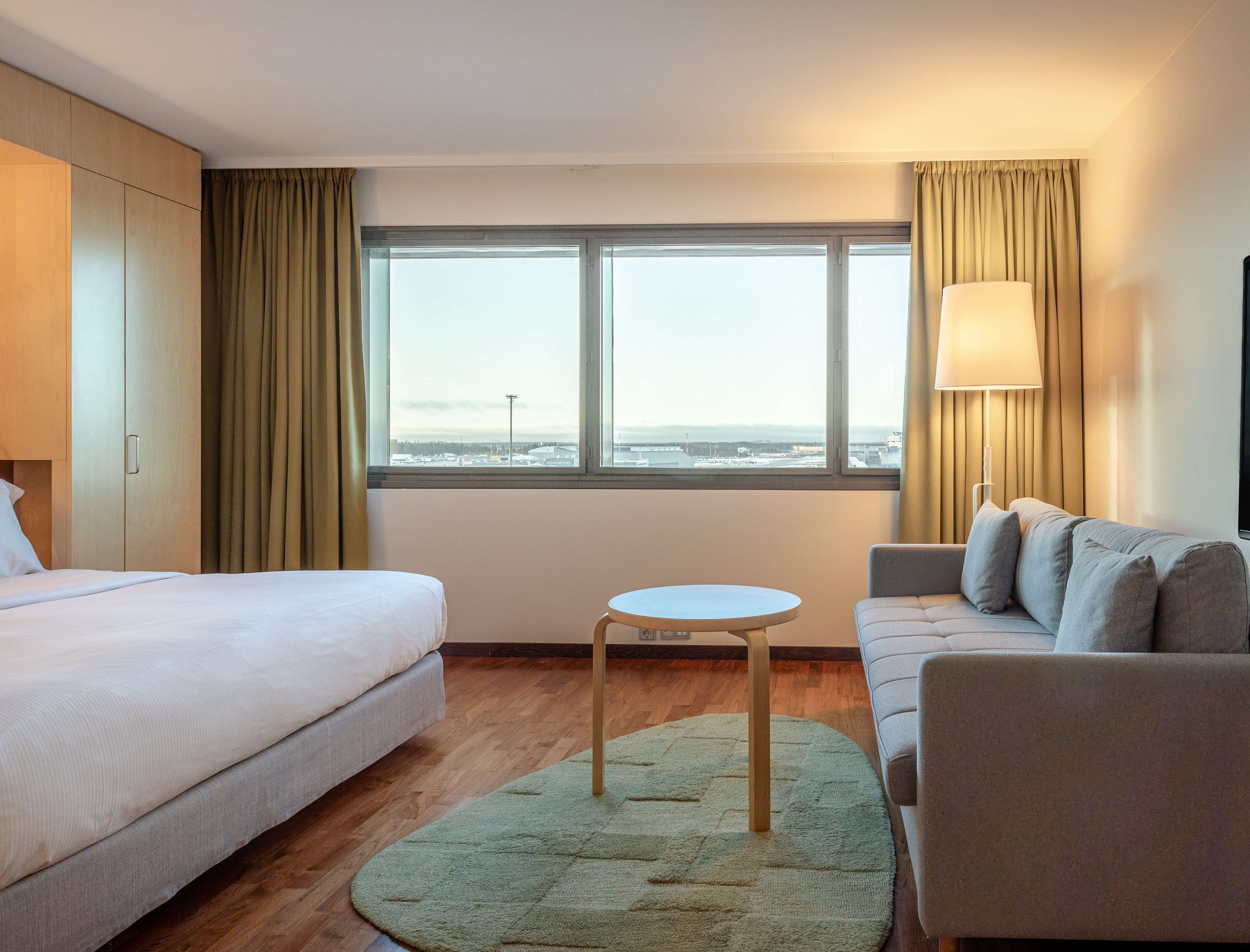 Habitació d'hotel amb llit, sofà, taula petita, làmpada i vista de la ciutat a través de grans finestres.