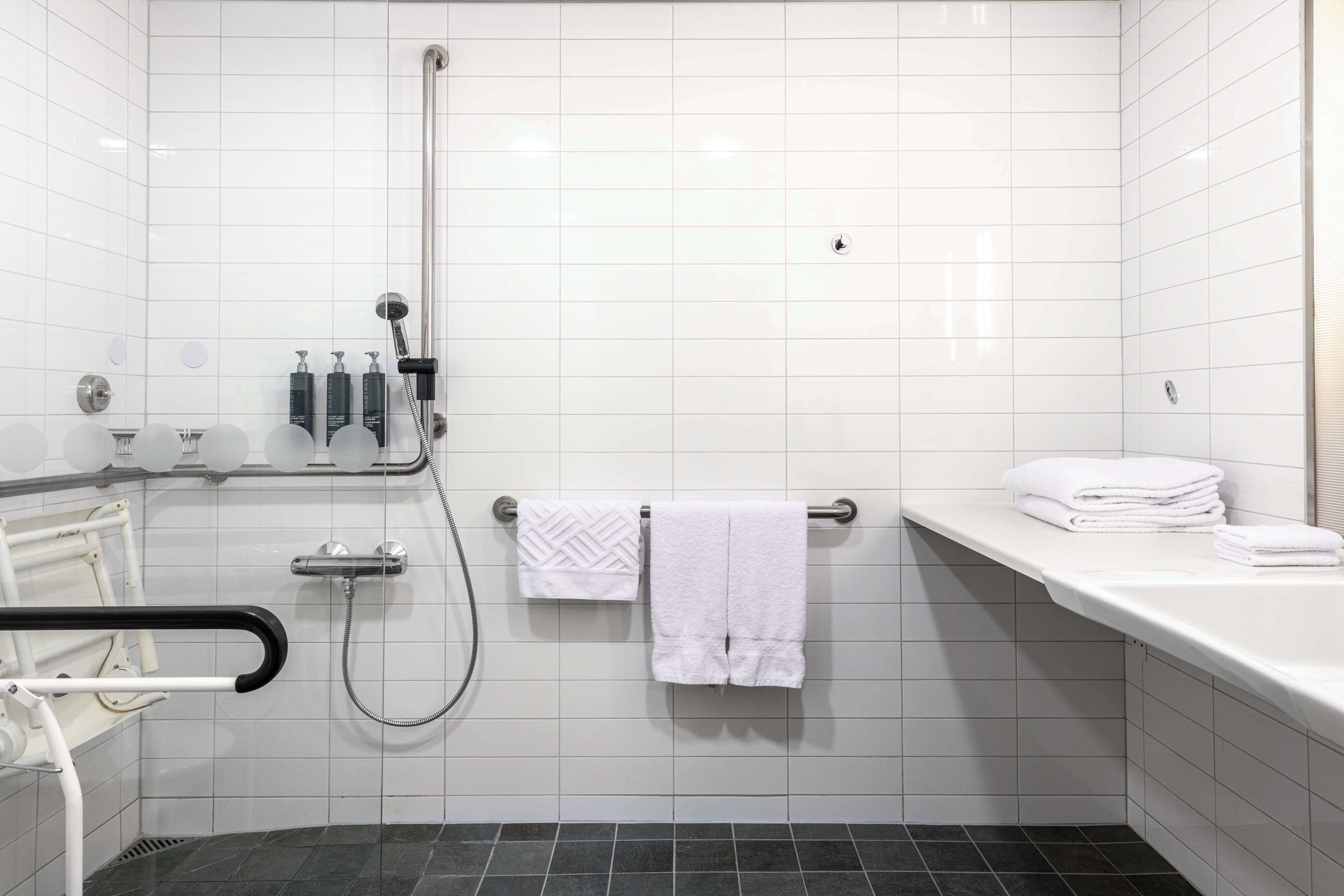Un baño moderno con azulejos blancos, zona de ducha, barra de apoyo y una pila de toallas blancas.