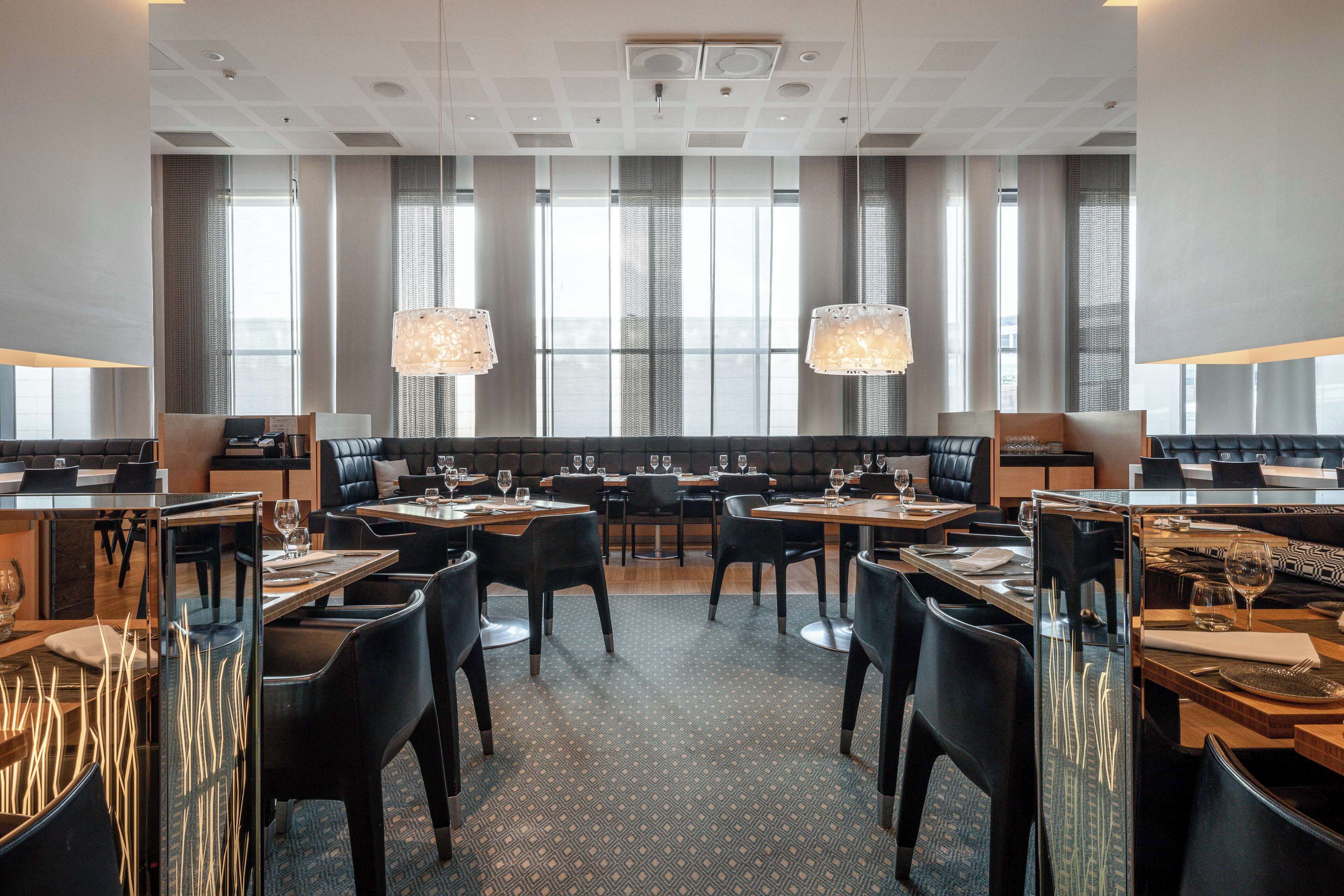 A modern dining area with multiple tables and chairs, large windows with curtains, and hanging pendant lights.