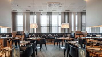 A modern dining area with multiple tables and chairs, large windows with curtains, and hanging pendant lights.