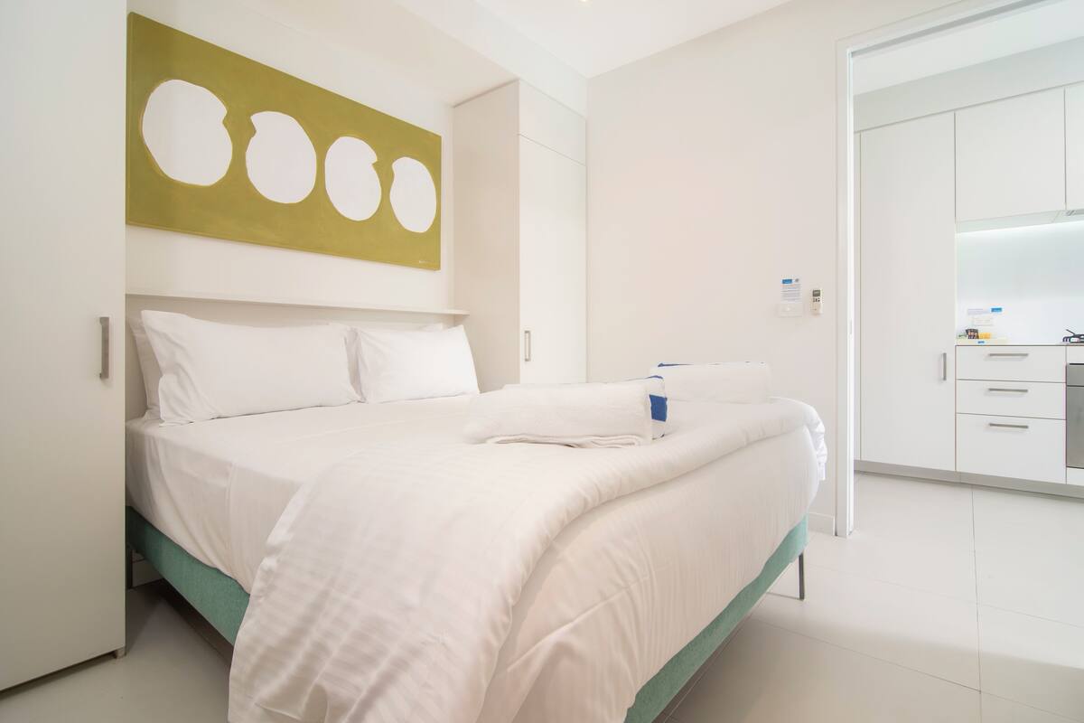A modern hotel room with a neatly made bed, a green accent on the bed, a large abstract painting on the wall, and a white wardrobe.