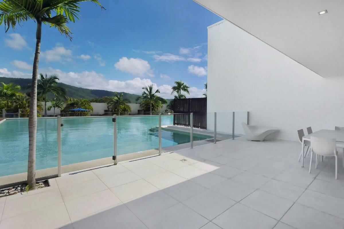 A modern outdoor pool area with a glass barrier, white furniture, and a view of mountains and palm trees.