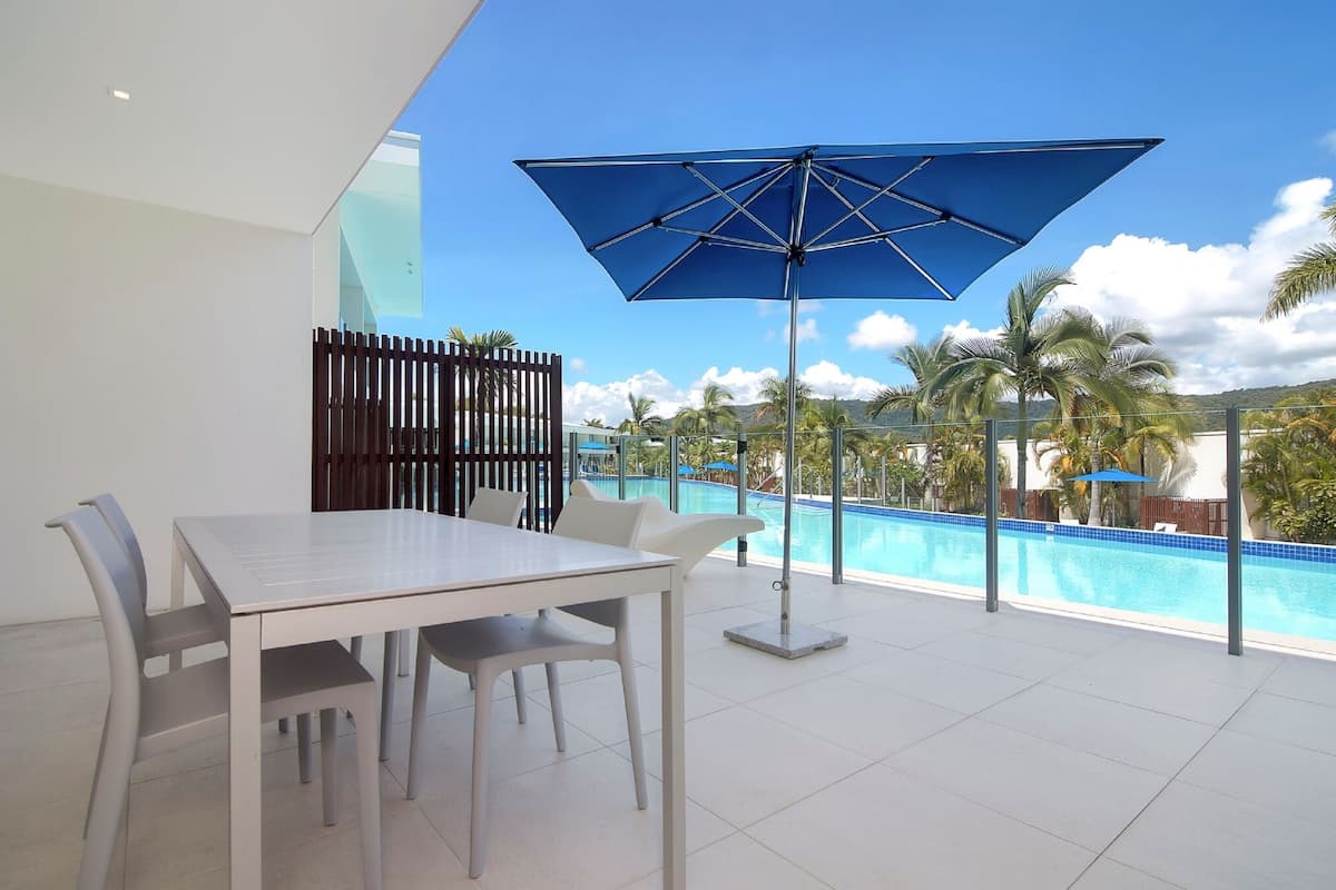 A modern outdoor patio with a pool, white table and chairs, and a blue umbrella.