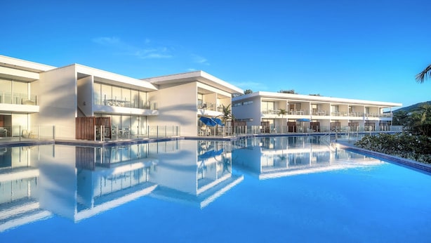 A modern hotel with a large swimming pool, multiple balconies, and a clear blue sky.