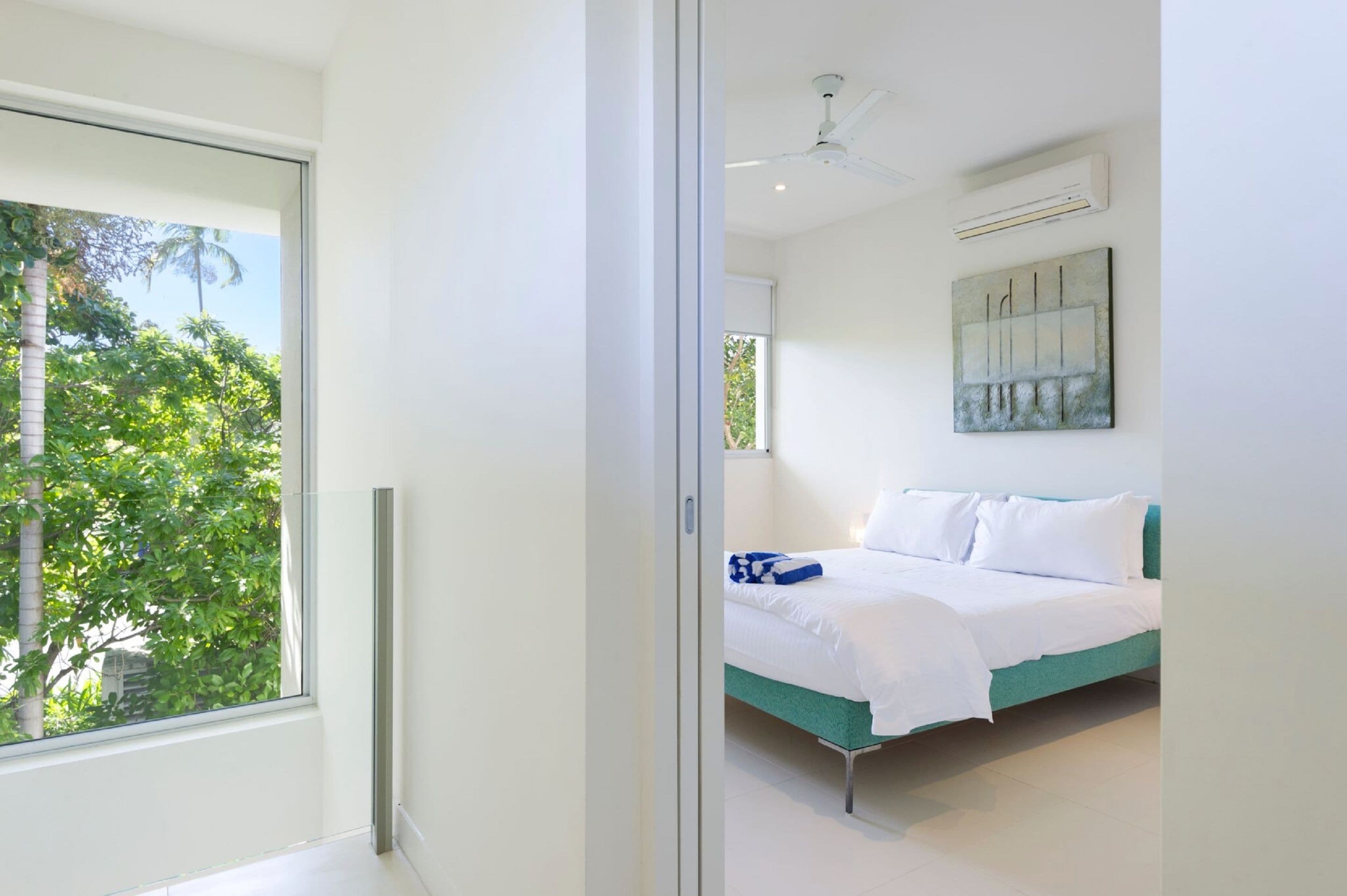 A modern bedroom with a large window, a bed with white linens, a teal accent wall, and a ceiling fan.