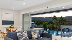 A modern living room with a sofa, a dining table, and a view of a swimming pool and mountains.