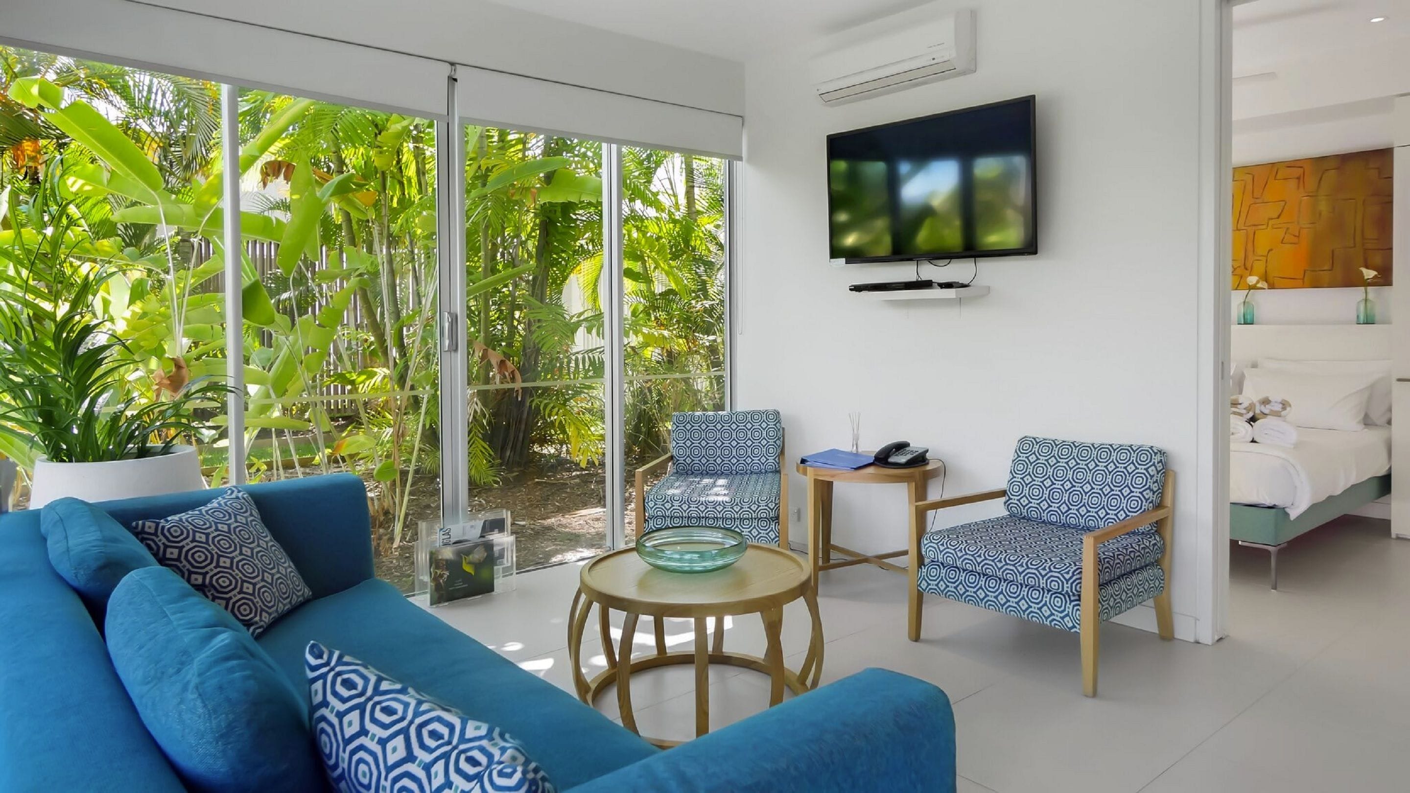 A modern living room with a blue sofa, two patterned armchairs, a glass coffee table, and a TV mounted on the wall.
