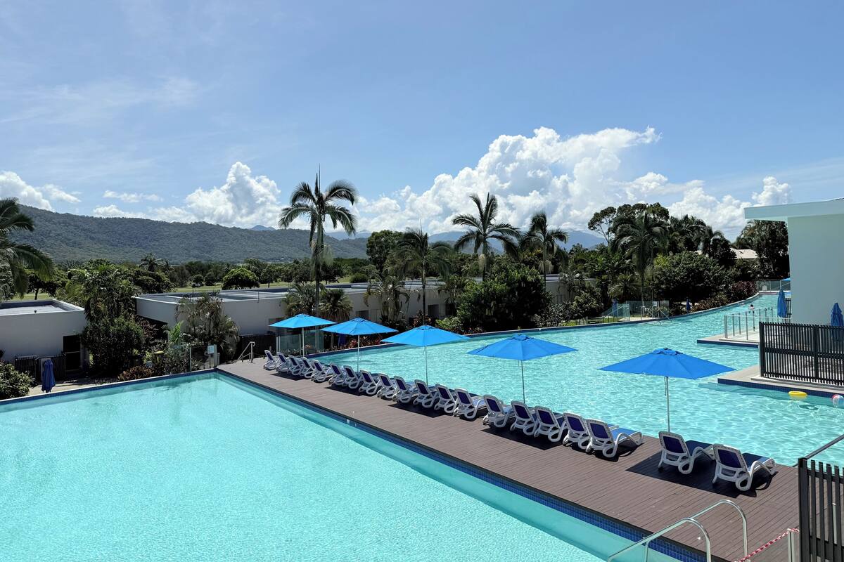 A swimming pool with lounge chairs and umbrellas, surrounded by palm trees and a mountainous backdrop.