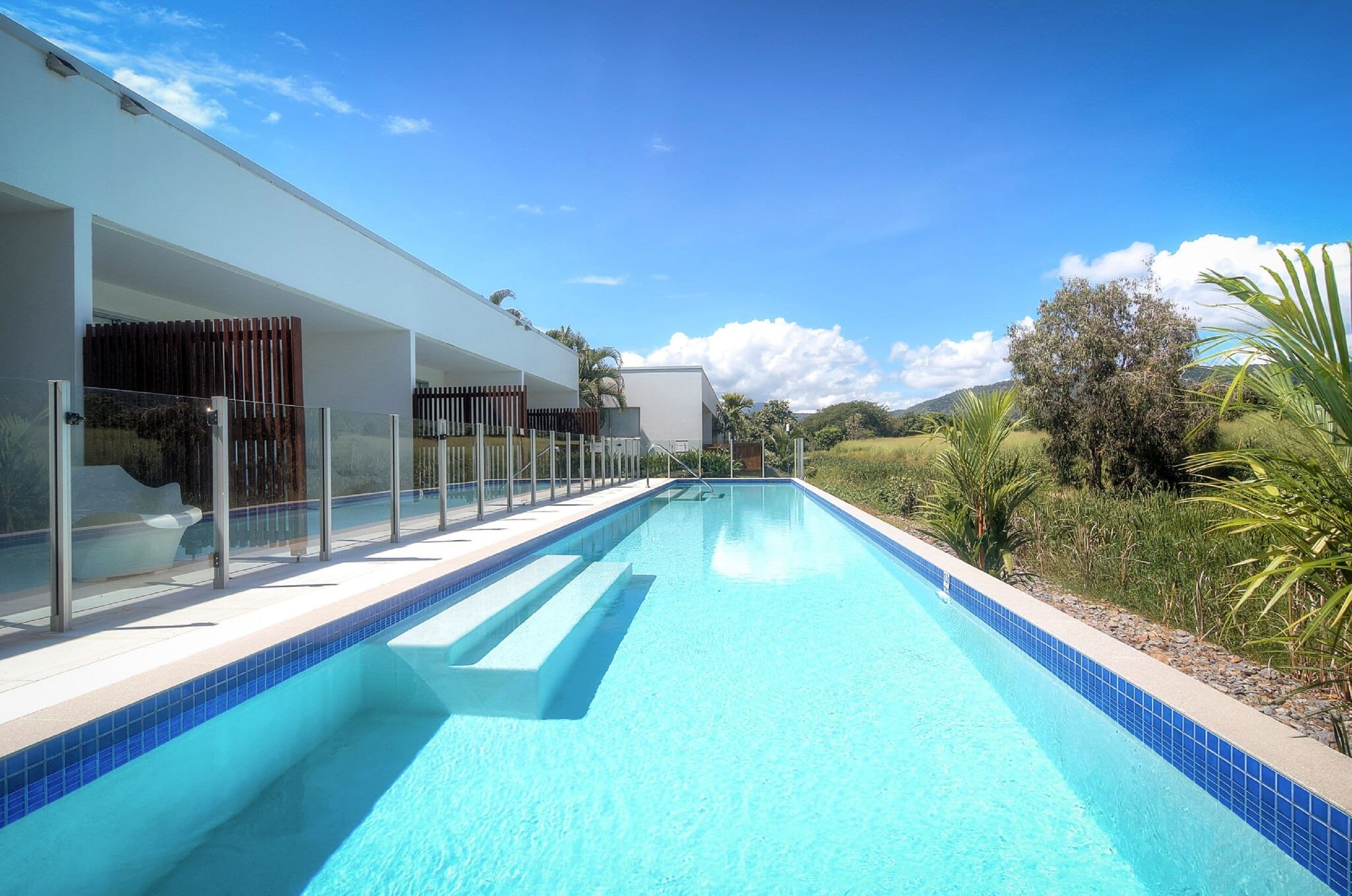 A modern pool area with clear blue water, surrounded by a white building with glass railings and a view of the countryside.