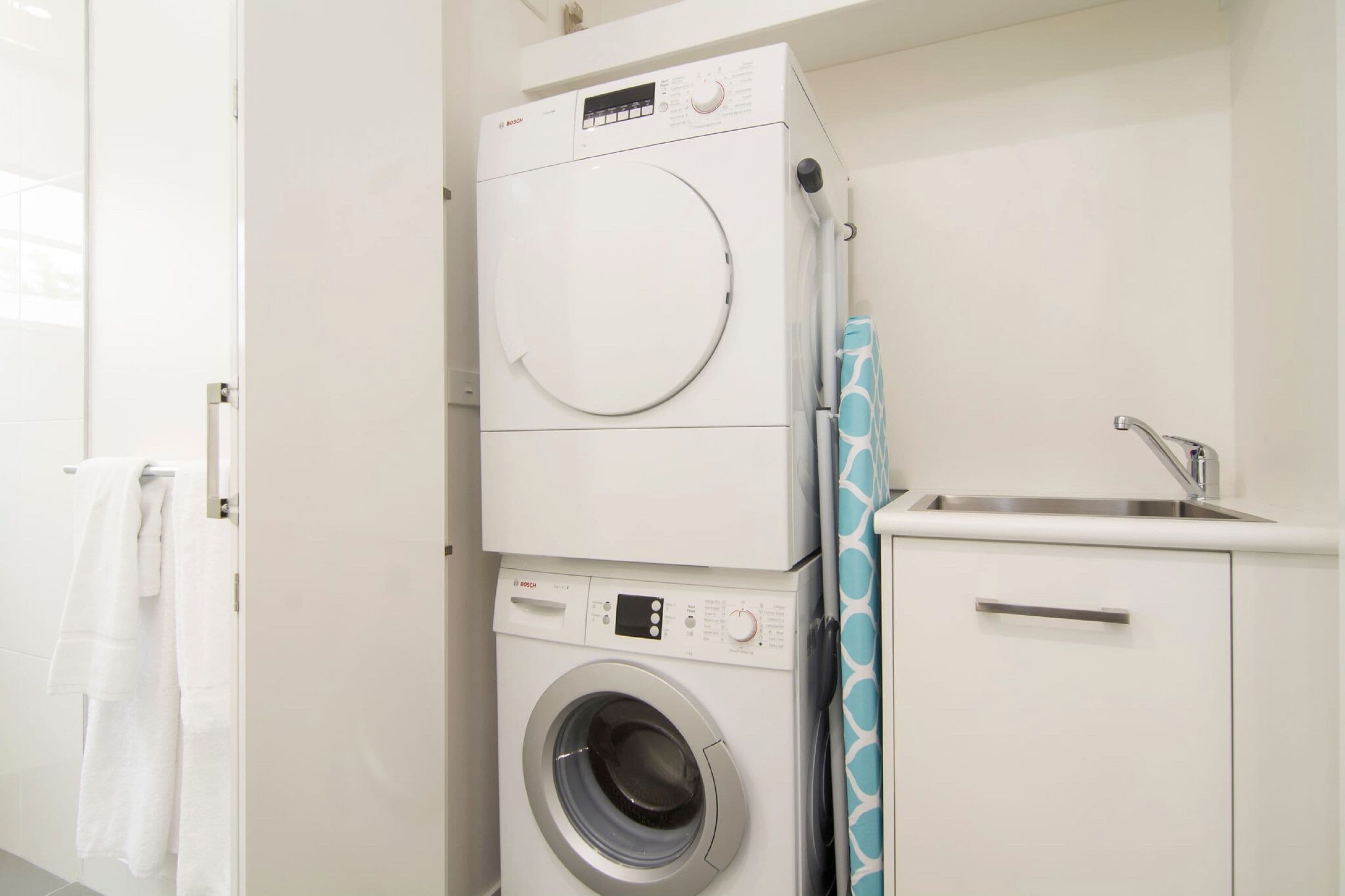 Een kleine wasruimte met een voorlader wasmachine en droger, een klein wastafeltje met kraan, en een handdoekrek.