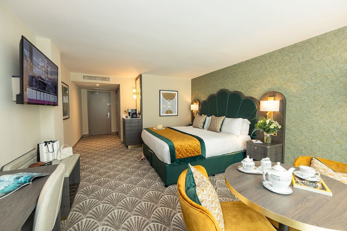 A hotel room with a bed, a desk, a chair, a table with tea set, and a TV on the wall.