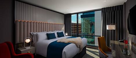 A modern hotel room with a large bed, a desk, a chair, and a view of a building through the window.