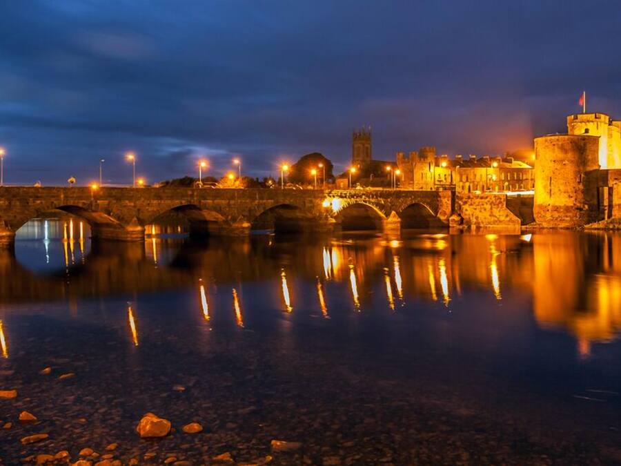 A stone bridge over a calm river at night, reflecting lights and buildings.