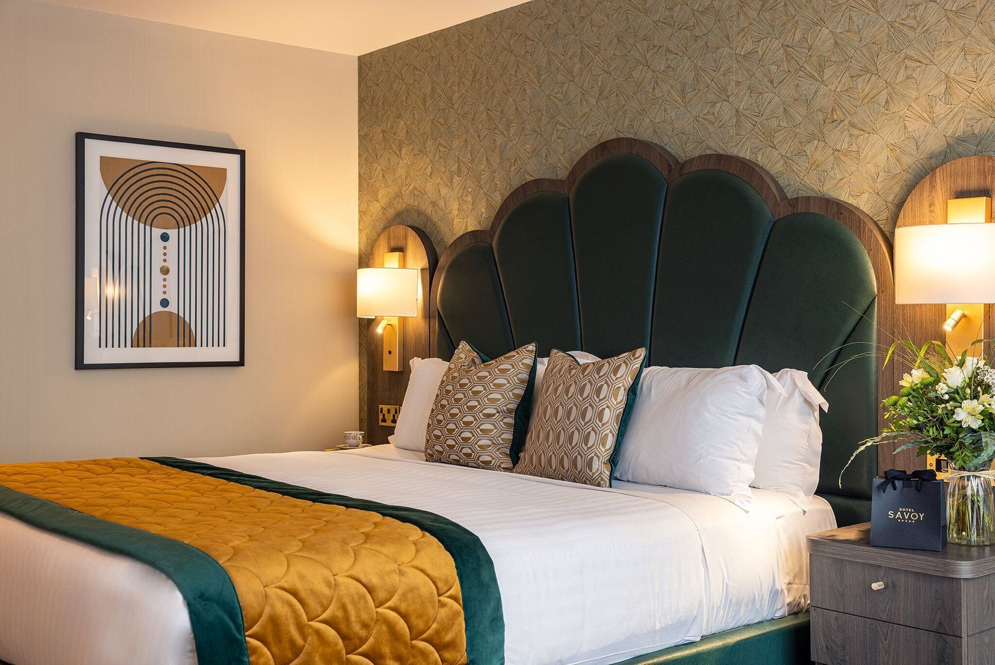 Un lit bien fait, avec une tête de lit verte, une literie blanche et un plaid de couleur moutarde. On y trouve des coussins à motifs, une œuvre d’art encadrée au-dessus du lit, ainsi qu’une table de chevet avec une lampe et un vase de fleurs.
