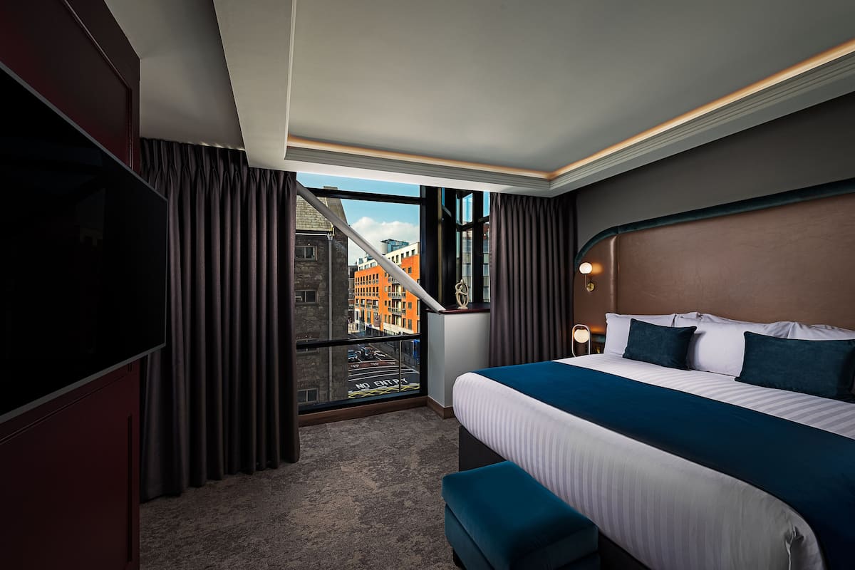 A hotel room with a large bed, a TV, a view of the city through a window, and a balcony.