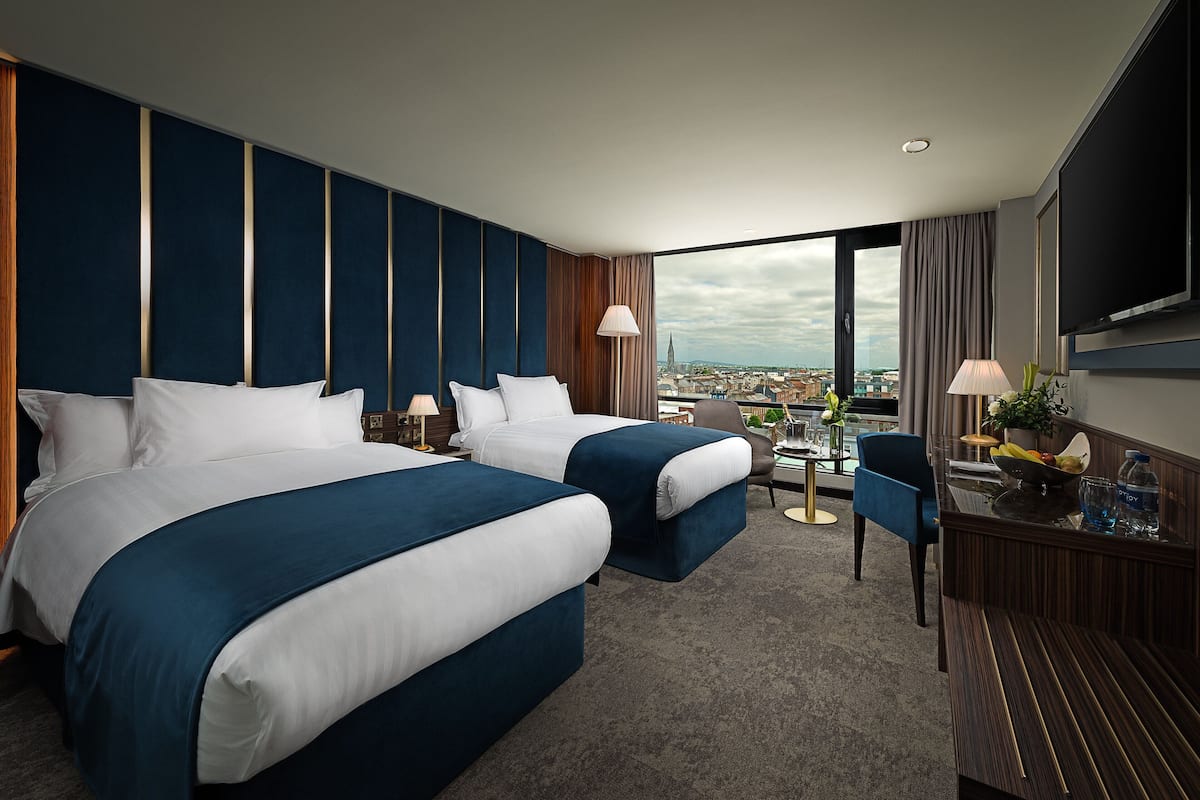 A hotel room with two beds, a desk, a chair, a TV, and a view of the city.