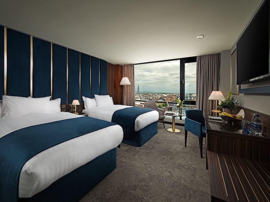 A hotel room with two beds, a desk, a chair, a TV, and a view of the city.