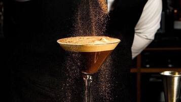 Un cantinero sirviendo una bebida en una copa de martini, sobre un fondo oscuro y una barra.