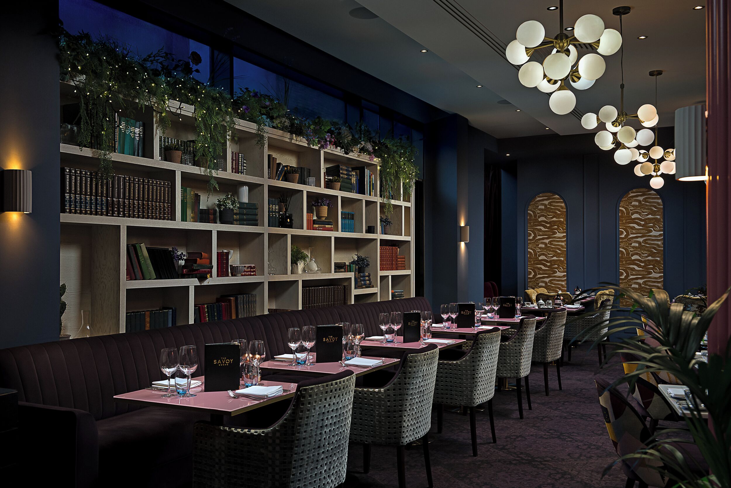A restaurant with a long table, cushioned seating, and a bookshelf with plants.