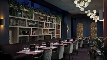 A restaurant with a long table, cushioned seating, and a bookshelf with plants.