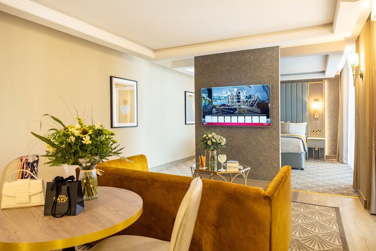 A hotel room with a sofa, a table with a vase of flowers, a TV on the wall, and a bed with a headboard.