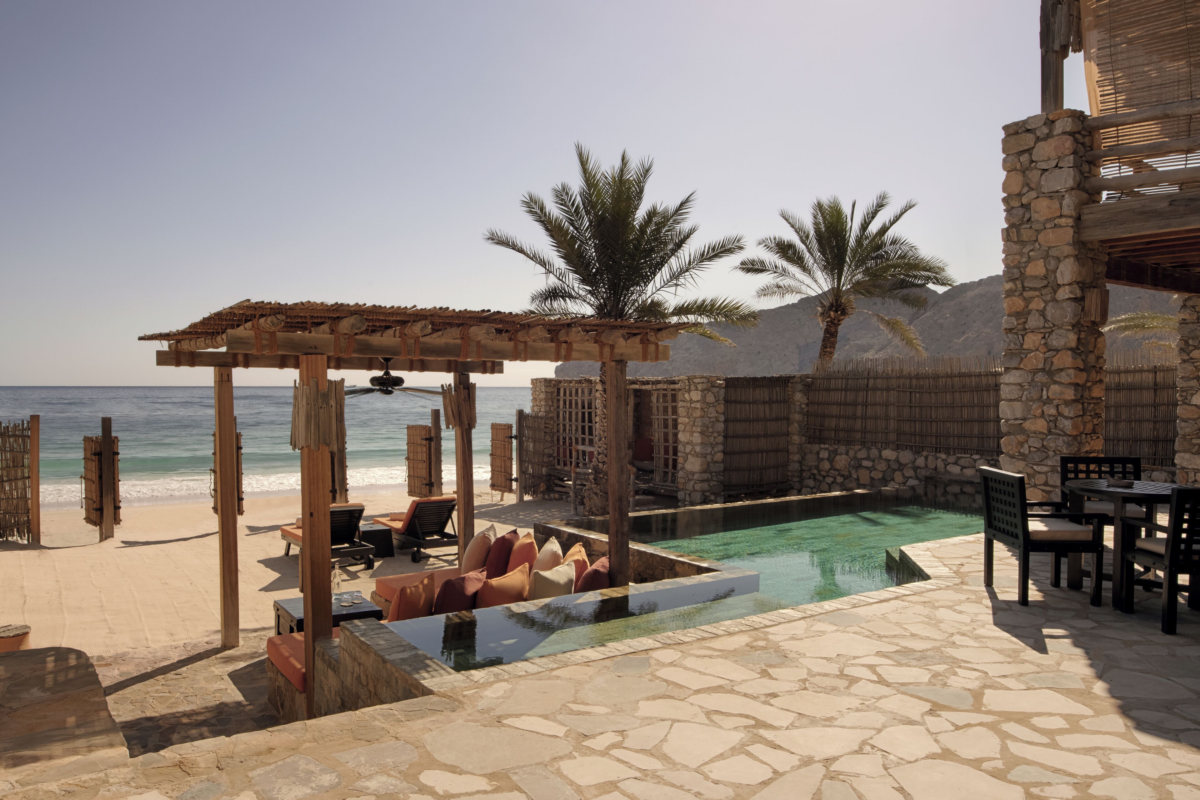 A pool area with stone flooring, lounge chairs, and a pergola overlooking the beach.