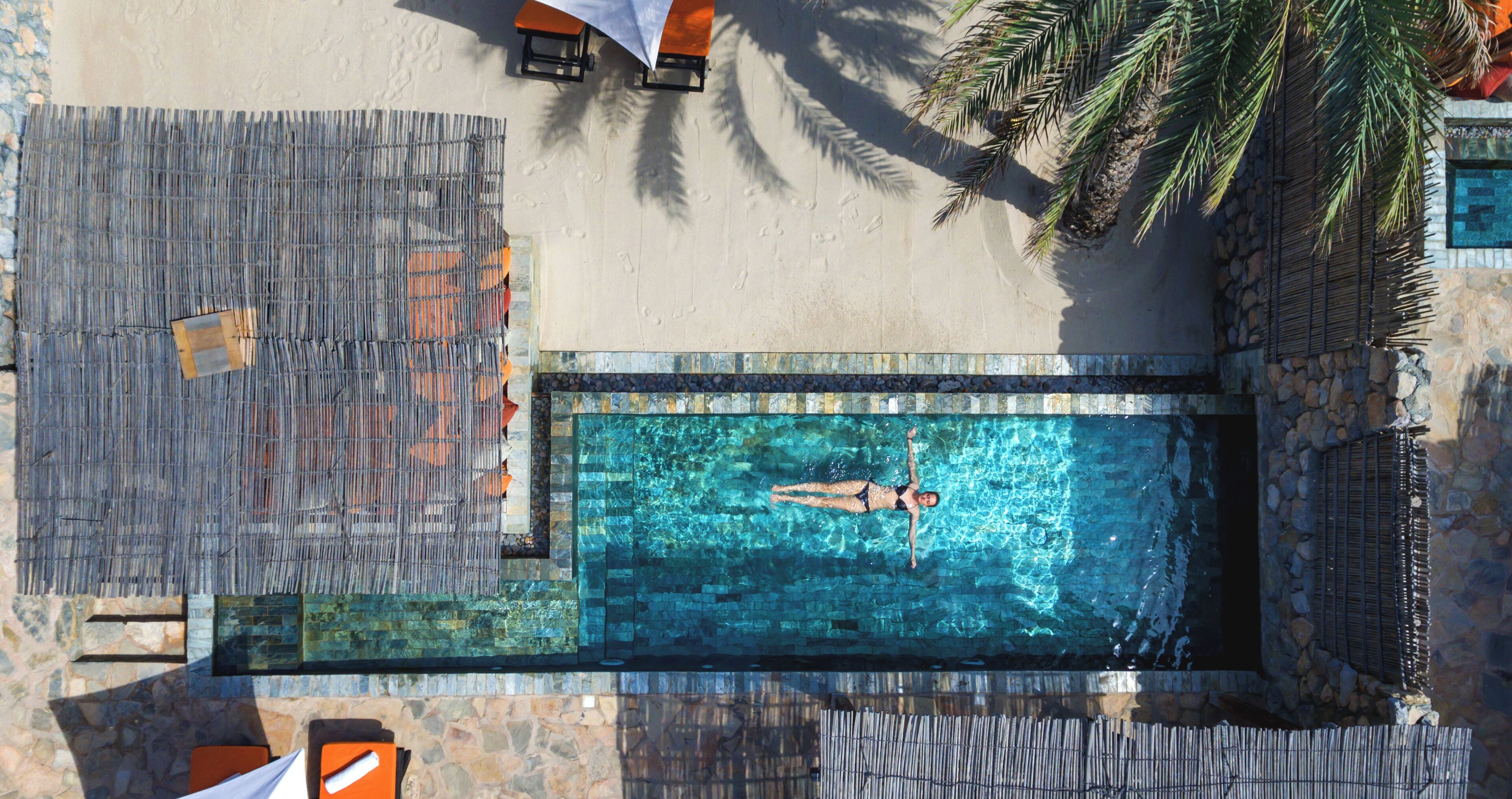 Vista aérea de una piscina con una persona nadando, rodeada de muros de piedra y palmeras.
