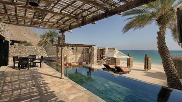 An infinity pool with a view of the sea, stone patio furniture, and a wooden pergola.