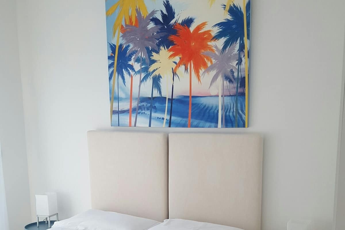 A bedroom with a large bed, a nightstand, a small table, and a painting of palm trees and a beach.