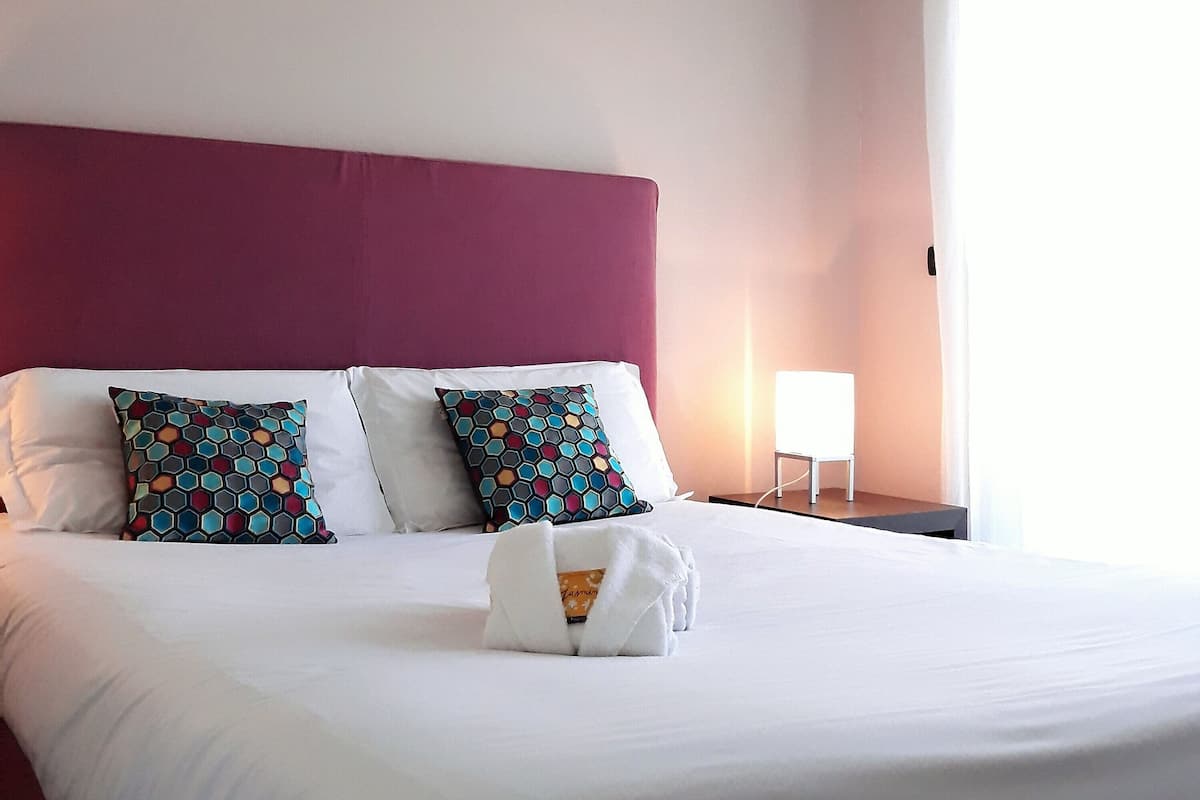 A hotel room with a large bed, two pillows, a bedside table with a lamp, and a window with curtains.