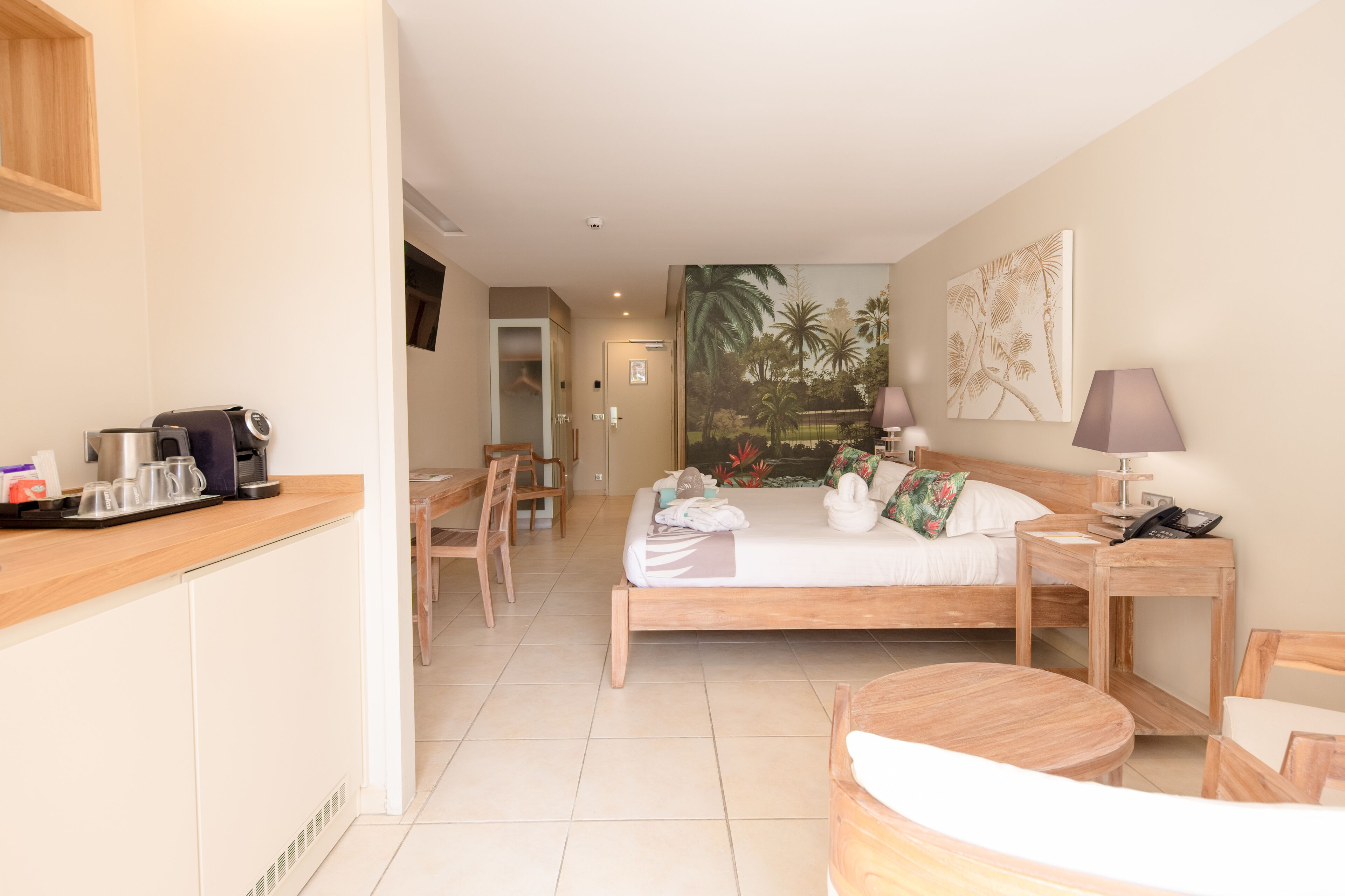Une chambre d’hôtel avec un lit, une table de chevet, une lampe et une petite table en bois. Il y a également un coin cuisine avec une machine à café et des tasses.