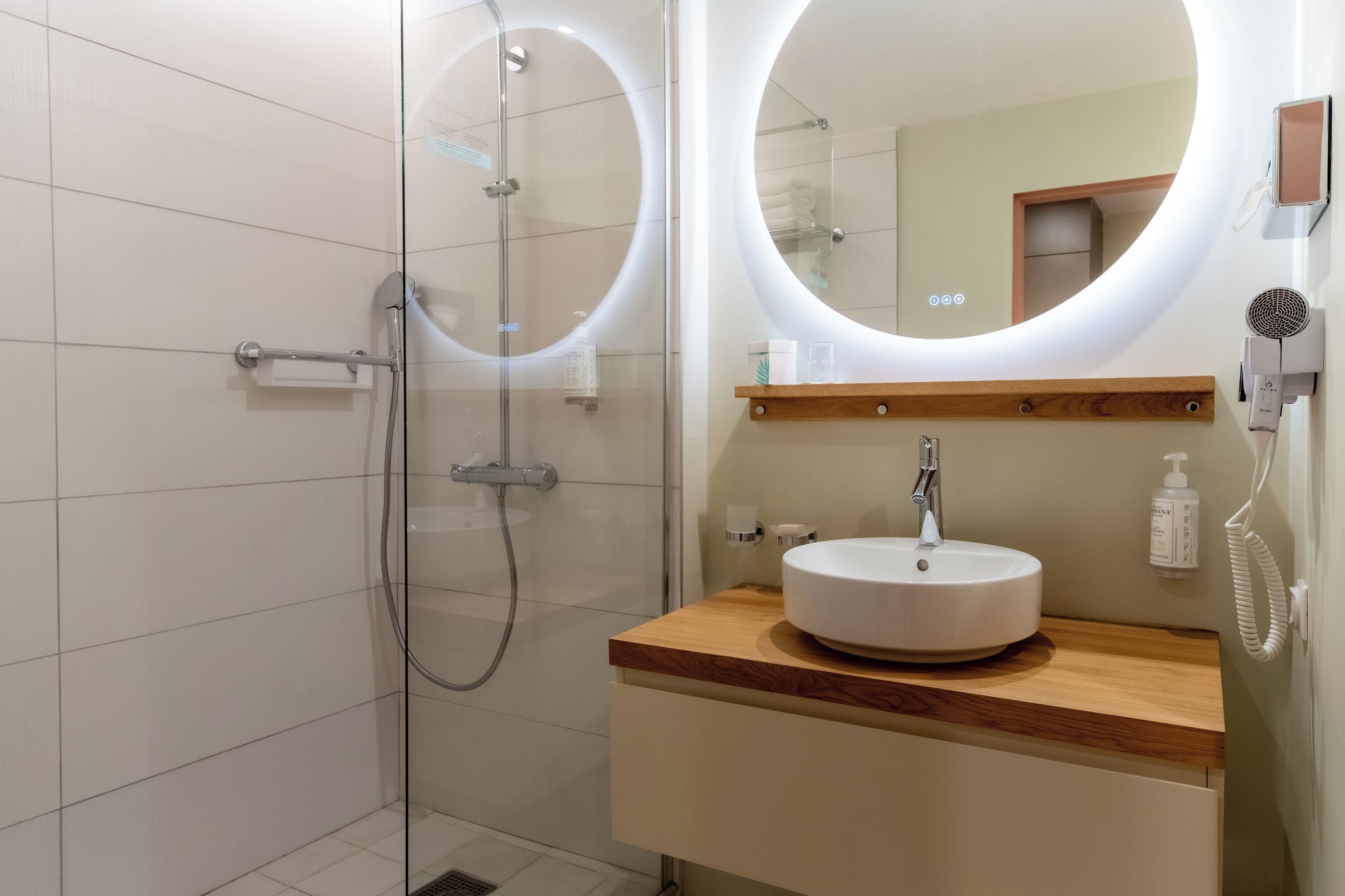 Un baño moderno con ducha de vidrio, tocador de madera con lavamanos blanco y un espejo grande con iluminación integrada.