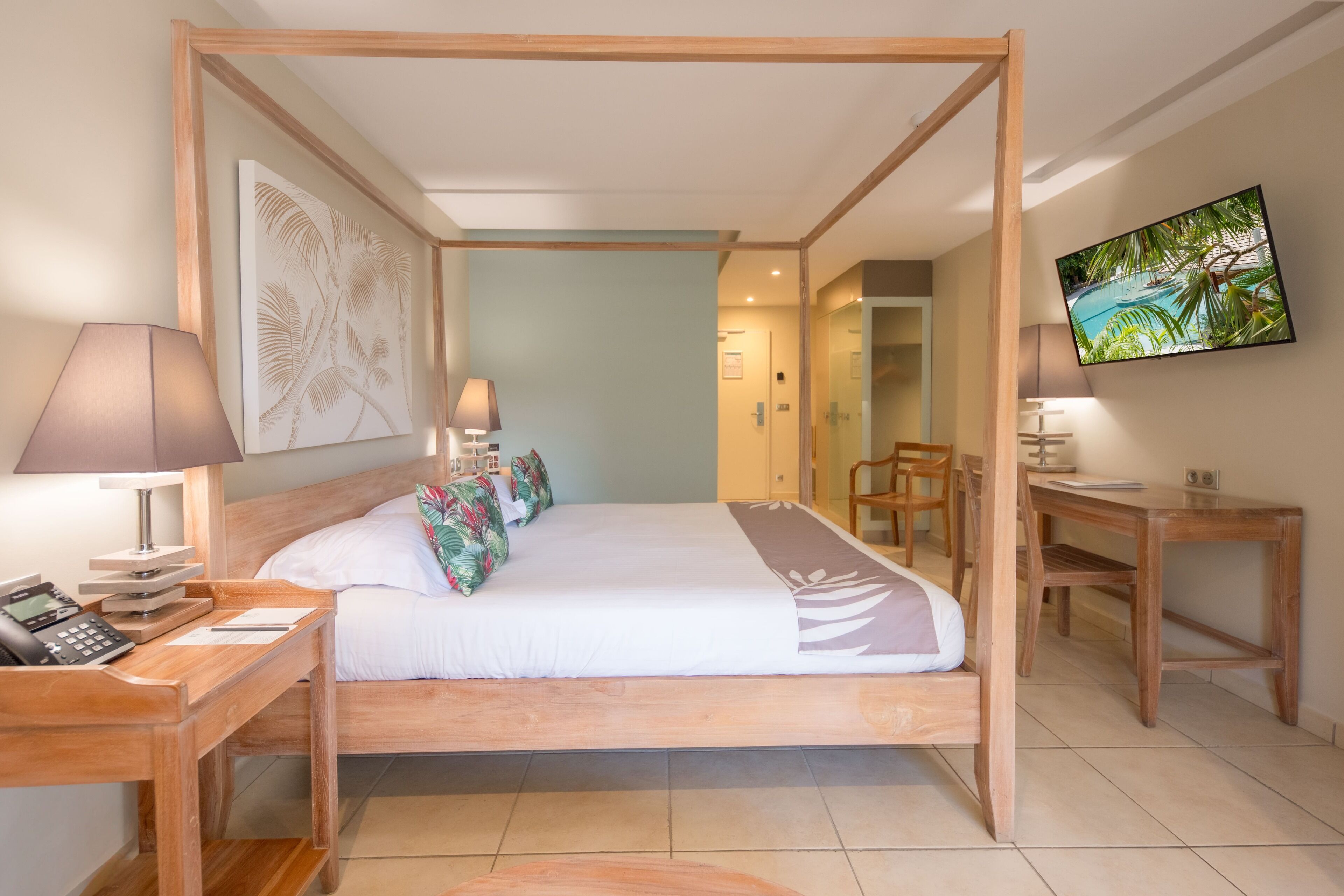 A four-poster bed with a wooden frame, a desk with a lamp, a telephone, and a TV on the wall.