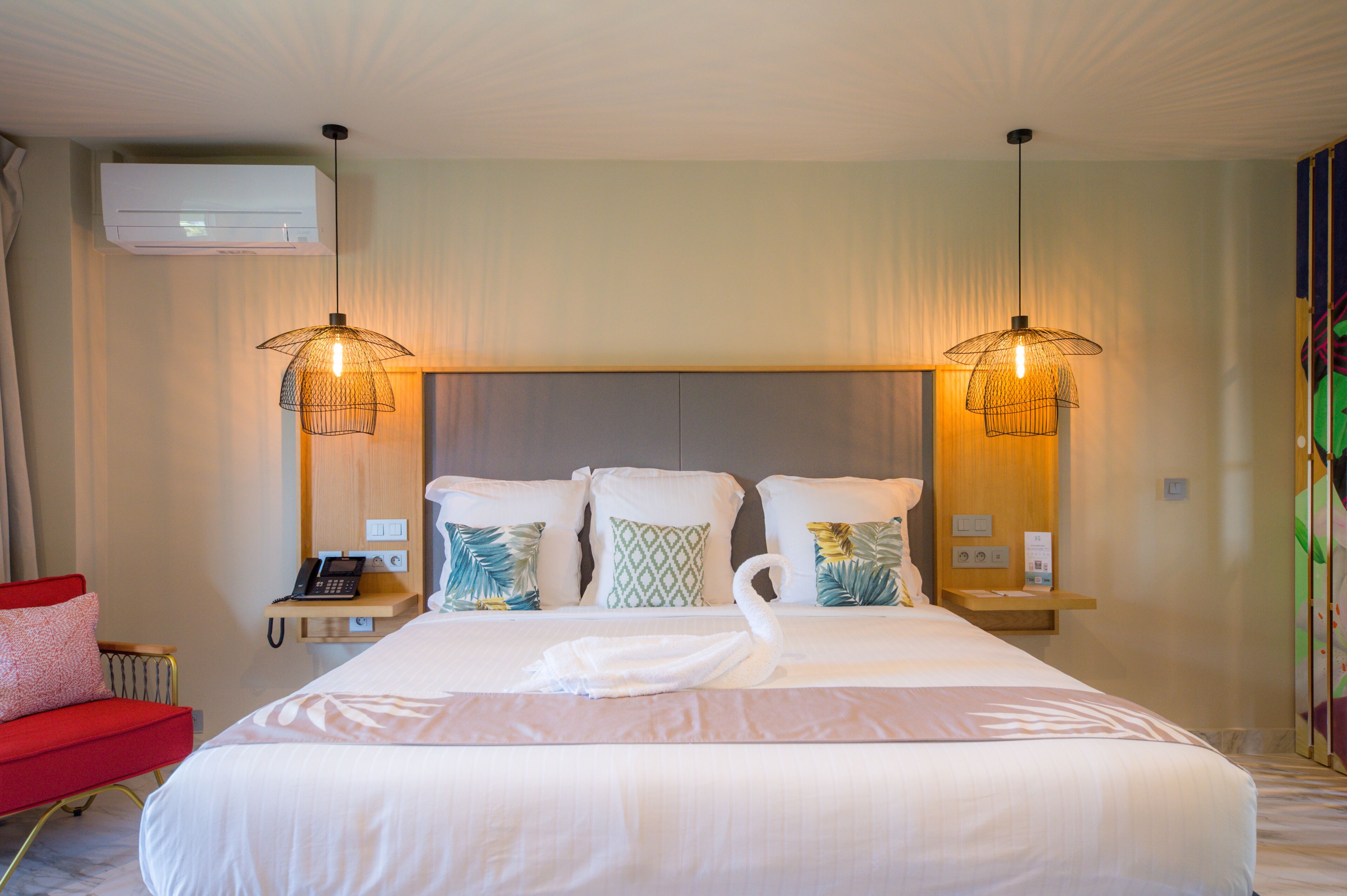 A hotel room with a large bed, two bedside tables, a red chair, and an air conditioning unit.