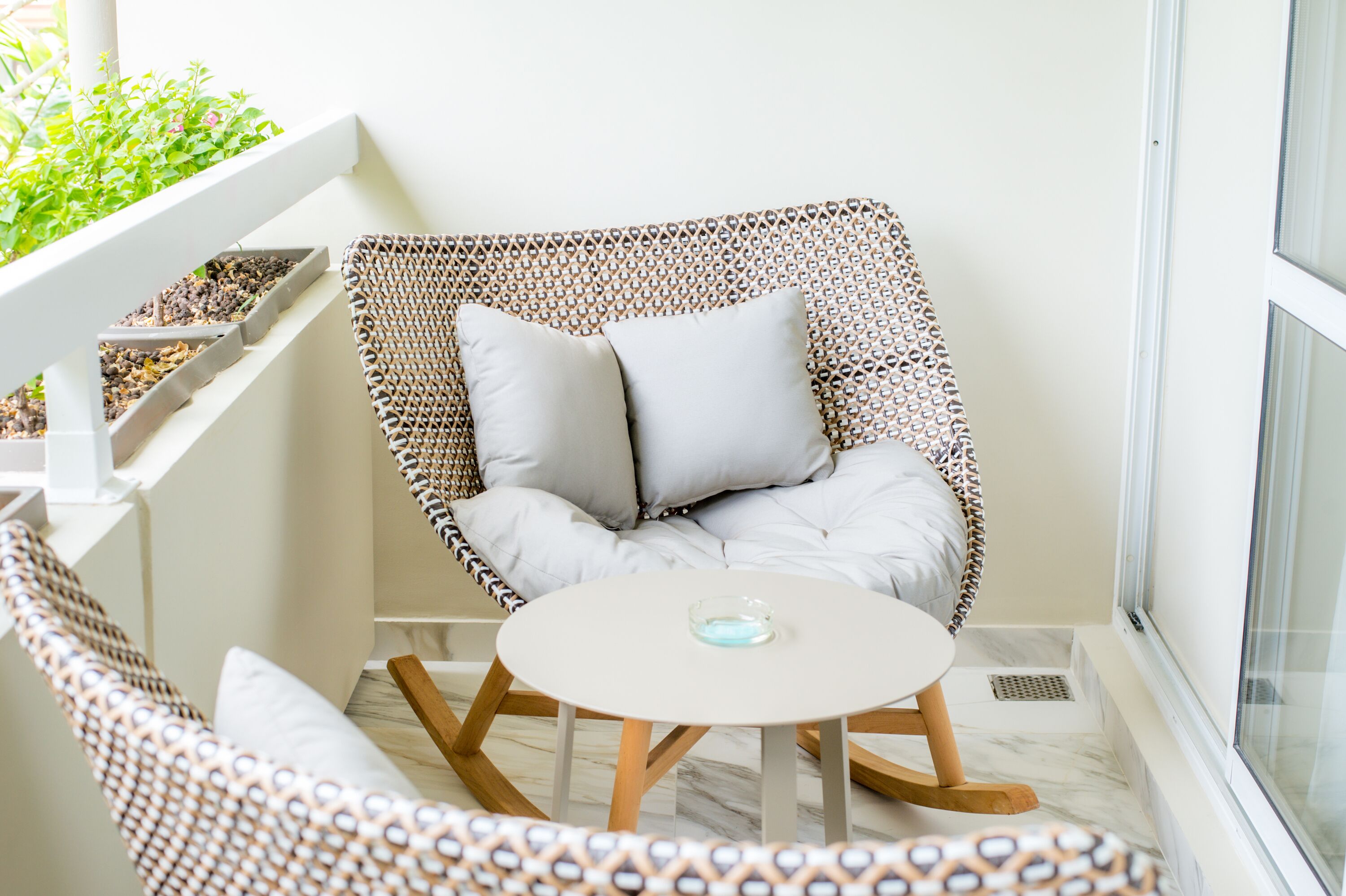 Une chaise en osier avec deux coussins, une petite table ronde sur laquelle se trouve un objet en verre, et une balustrade blanche agrémentée d’un jardinière.