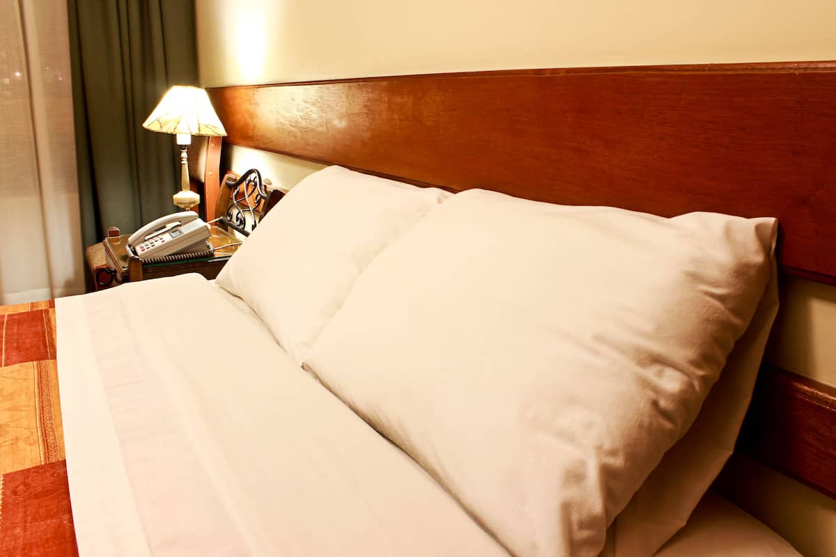 Un lit bien fait, avec du linge de lit blanc, une tête de lit en bois, une lampe de chevet et un téléphone posé sur une table de chevet en bois.